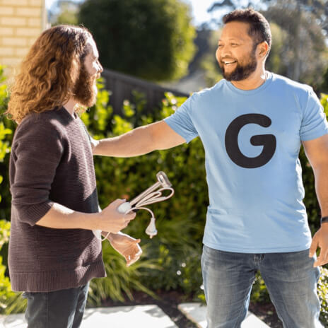 Two men greeting each other outside. One man with long hair holds cable cords, while the other, wearing a blue G shirt, smiles.